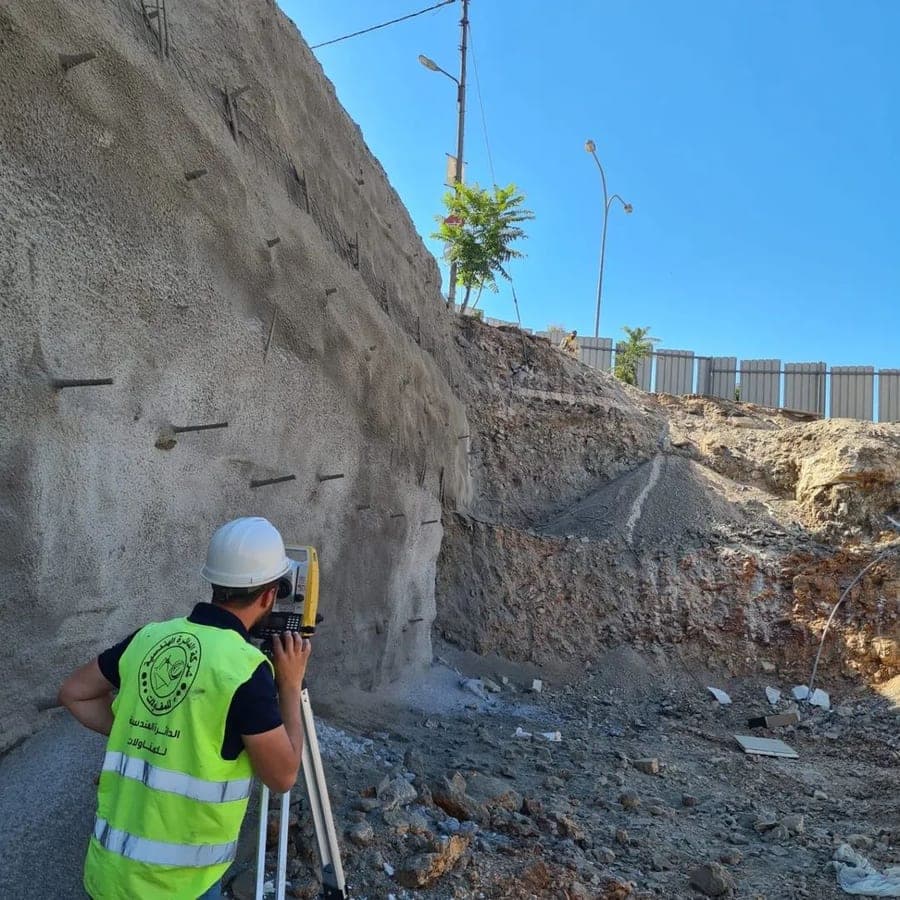 مسّاحٌ بسترة الشركة وخوذة يرصد بجهاز توتال ستيشن من توبكون (Topcon) أمام جدار حفريةٍ مدعَّمٍ بالشورنج